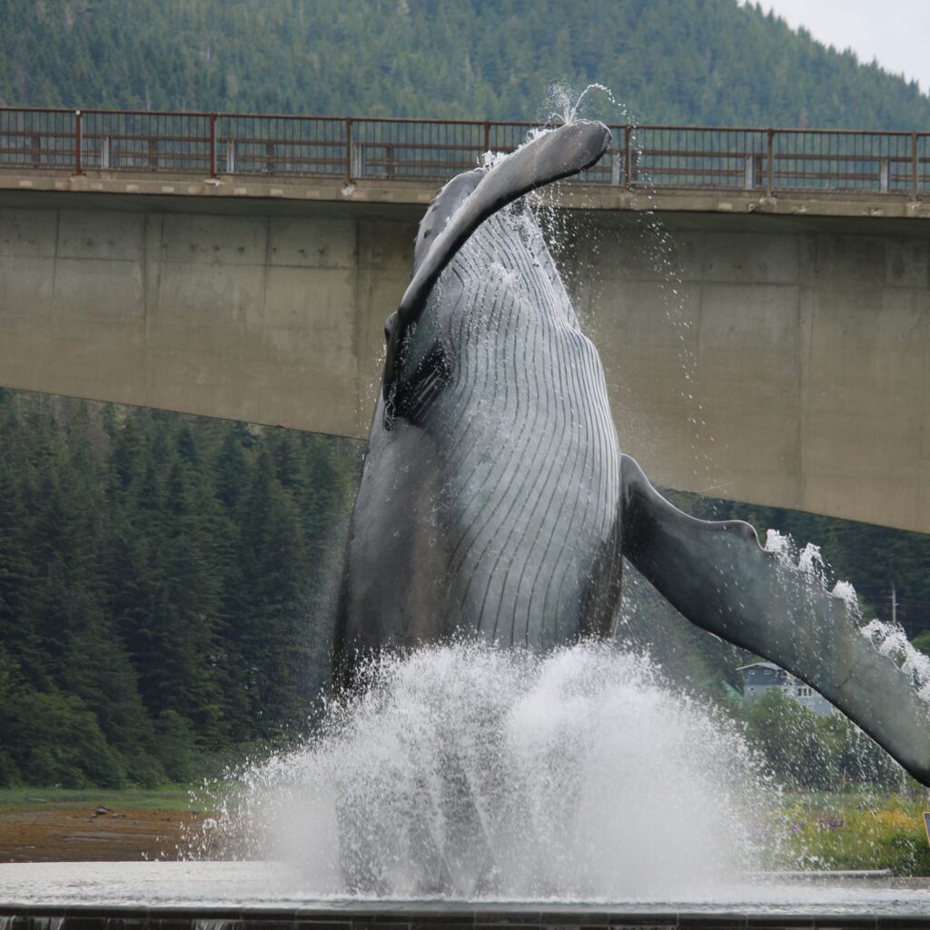 A large whale statue, breaching from the water, is set against a backdrop of forested mountains and a bridge. Water splashes around the base of the sculpture.