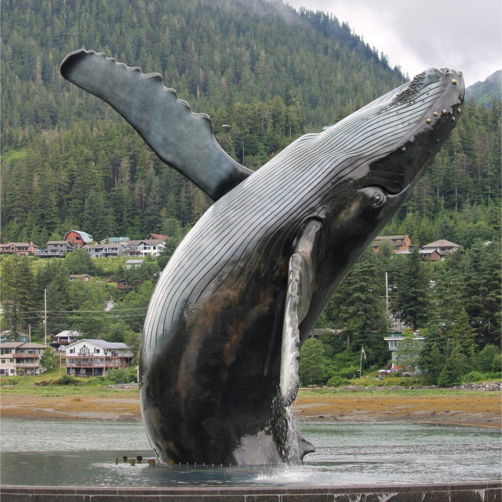 A large whale statue, breaching from the water, is set against a backdrop of forested mountains and hillside homes. Water splashes around the base of the sculpture.