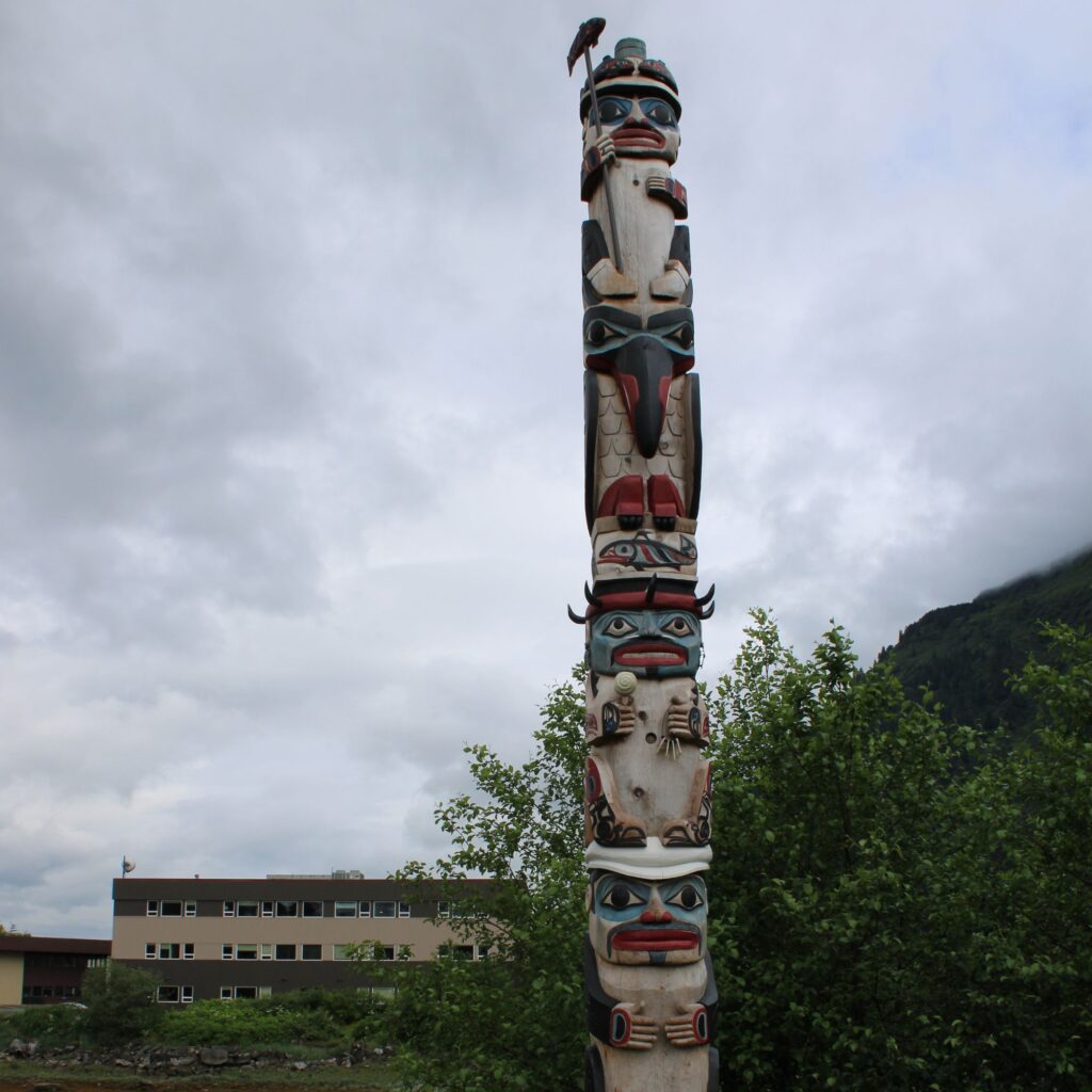 A tall, carved wooden totem pole featuring stacked figures painted in red, black, and blue, standing outdoors against a cloudy sky, am three story building with windows and green trees.
