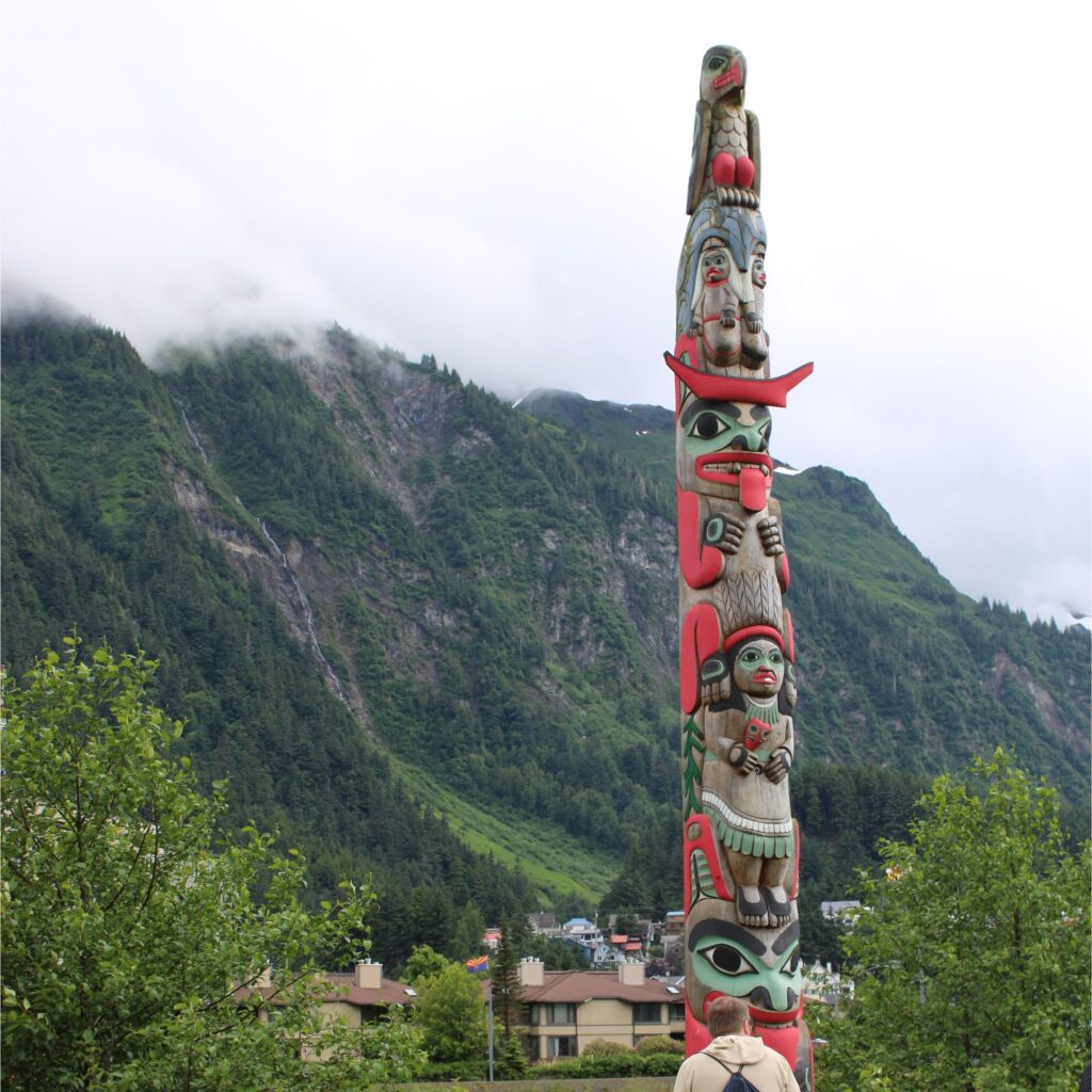A tall, carved wooden totem pole featuring stacked figures painted in red, black, and green, standing outdoors against a cloudy sky, mountains and green trees.