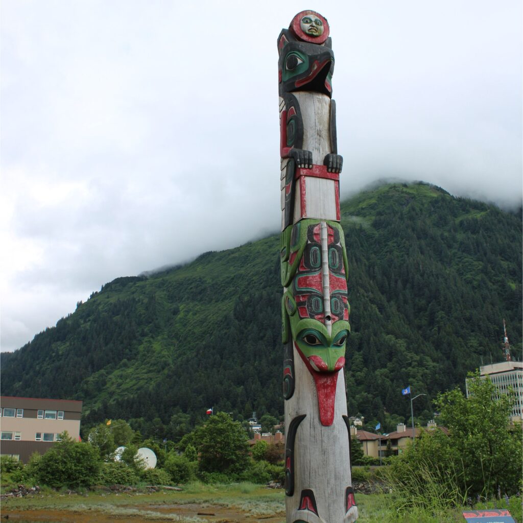 A tall, carved wooden totem pole featuring stacked figures painted in red, black, green, and blue, standing outdoors against a cloudy sky and green mountains.