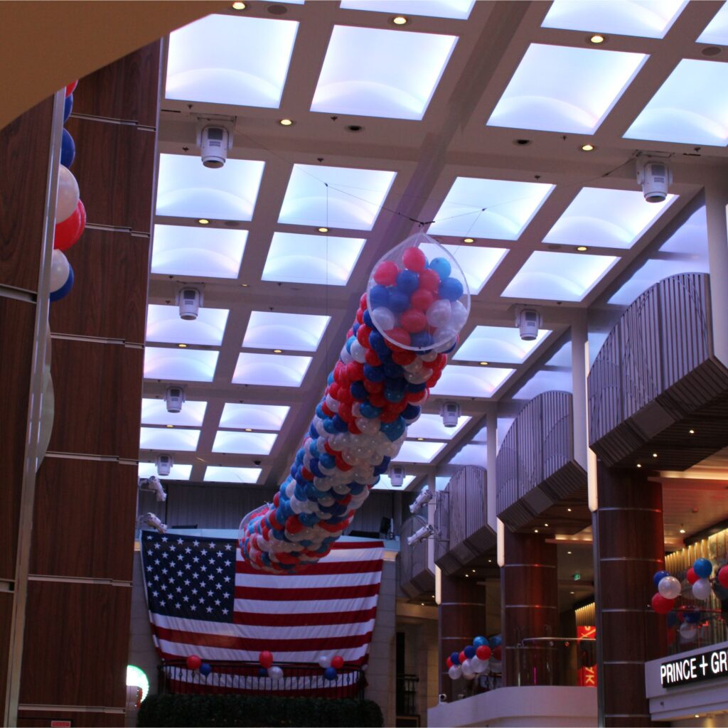 An interior space with a high ceiling, featuring an American flag made of balloons, and decorative blue, red, and white balloons hanging from the ceiling.