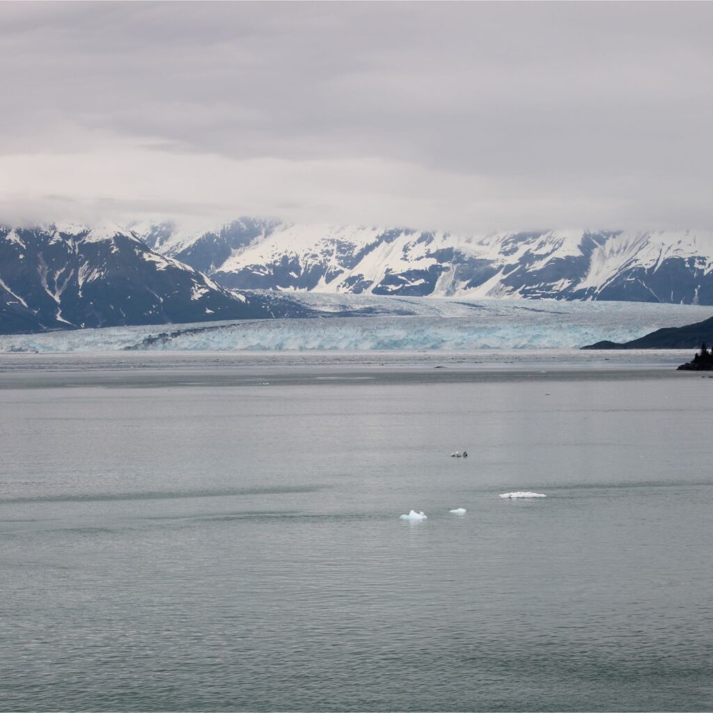 A serene landscape featuring a body of water in the foreground, with glaciers and snow-capped mountains in the background under a cloudy sky.
