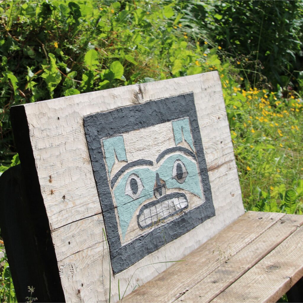 A wooden bench with a totem pole face on it that is surrounded by greenery.