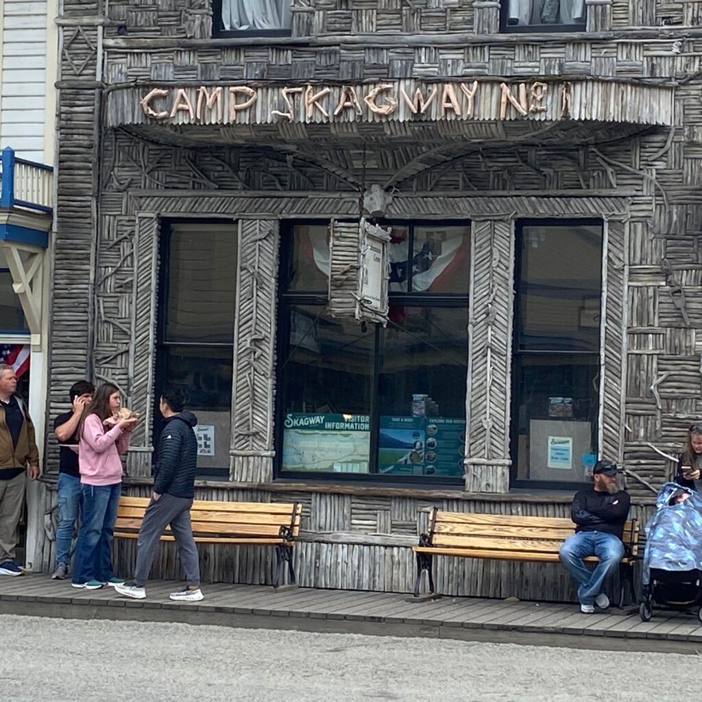 A wooden building  that says Camp Skagway No. 1 with large windows and benches out front with people sitting on them.