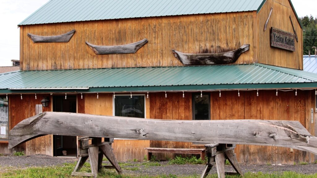 A wooden building with a green metal roof, featuring several wooden boat displays on the upper part of the facade and a large log positioned in front, resting on two supports.