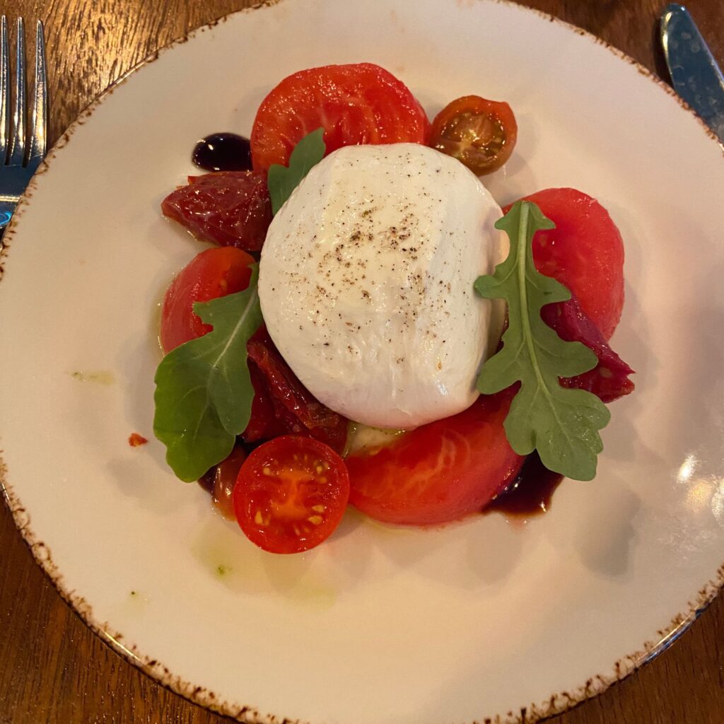 A plate of fresh tomatoes, including sliced red and cherry tomatoes, topped with a creamy burrata cheese and garnished with arugula. 