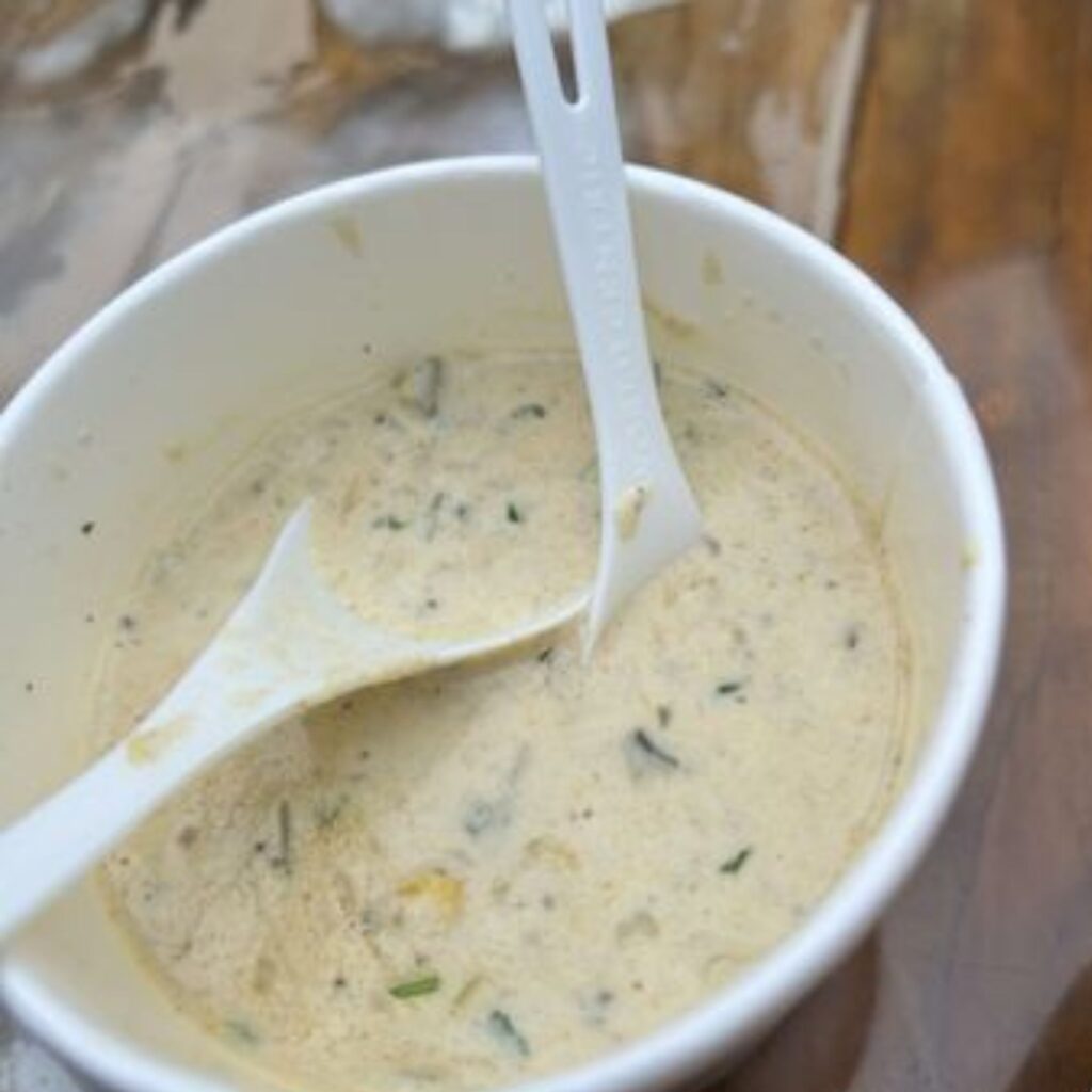 A white paper bowl contains creamy soup with herbs and two white plastic spoons.