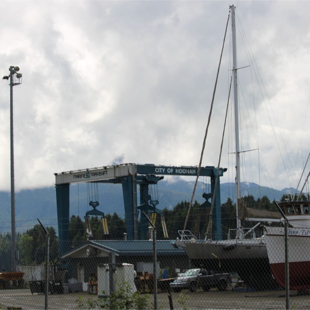 A boatyard with a large crane in the foreground, mountains in the background, and a cloudy sky.