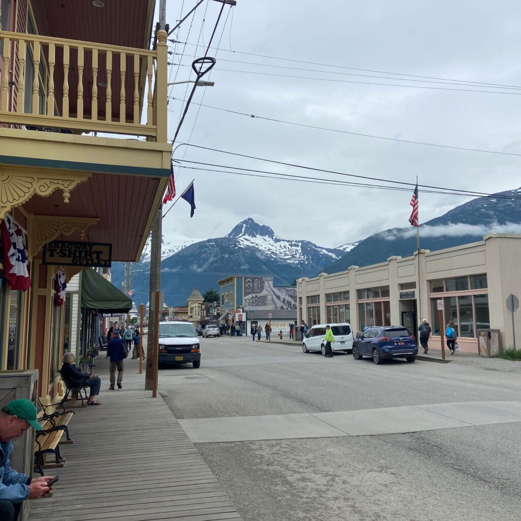 A street view of a small town with wooden buildings, people walking, and a mountain in the background partially covered with snow.