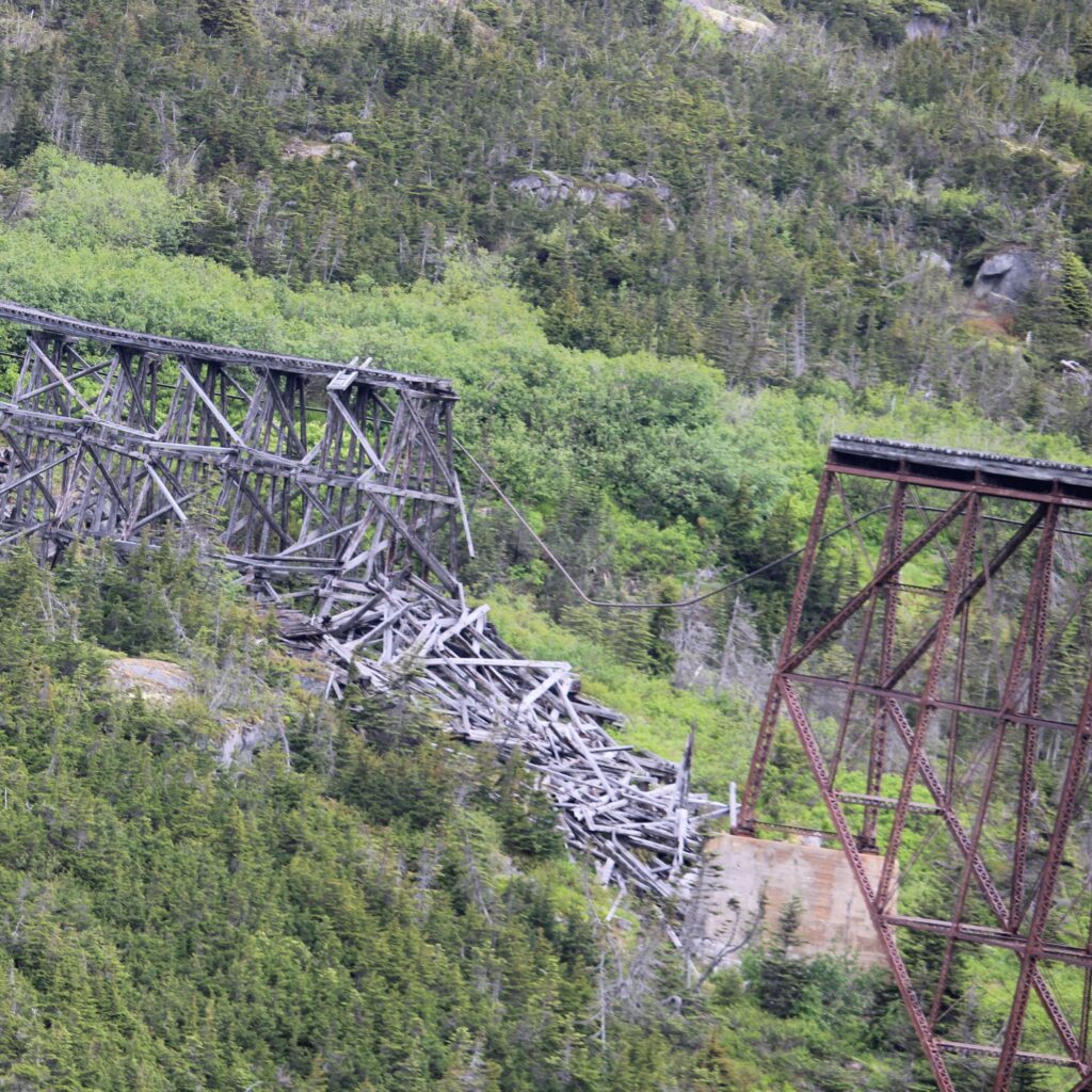 A dilapidated wooden bridge surrounded by dense greenery and mountains.