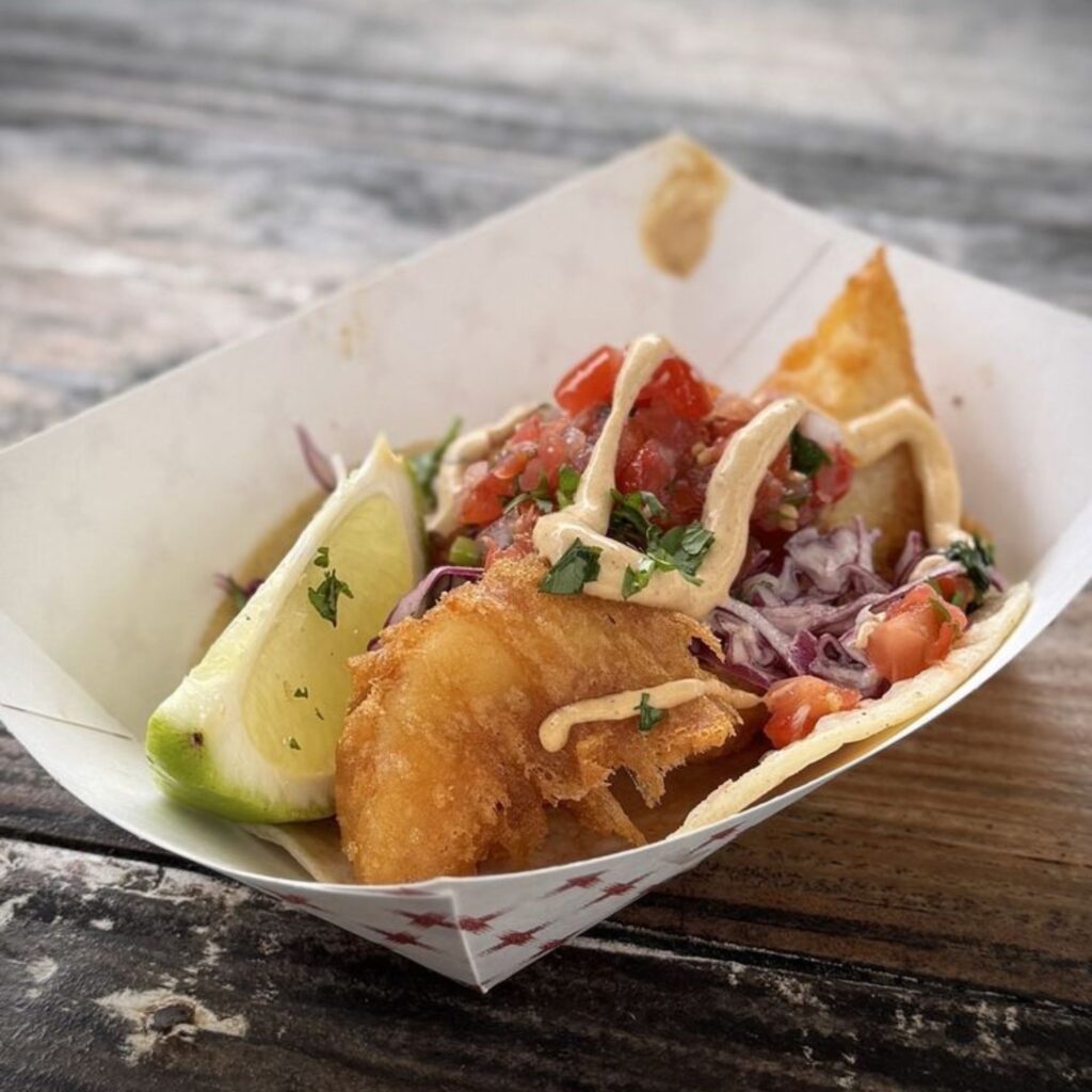 A fish taco in a paper tray, topped with slaw, salsa, creamy sauce, and cilantro, served with a lime wedge on a wooden surface.