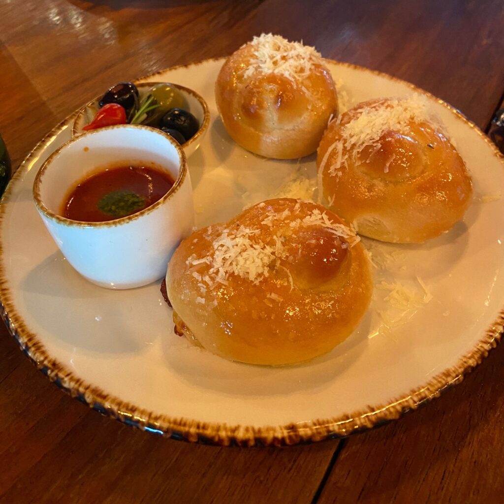 A plate with three golden-brown baked rolls sprinkled with cheese, accompanied by a small cup of dipping sauce and garnished with olives.

