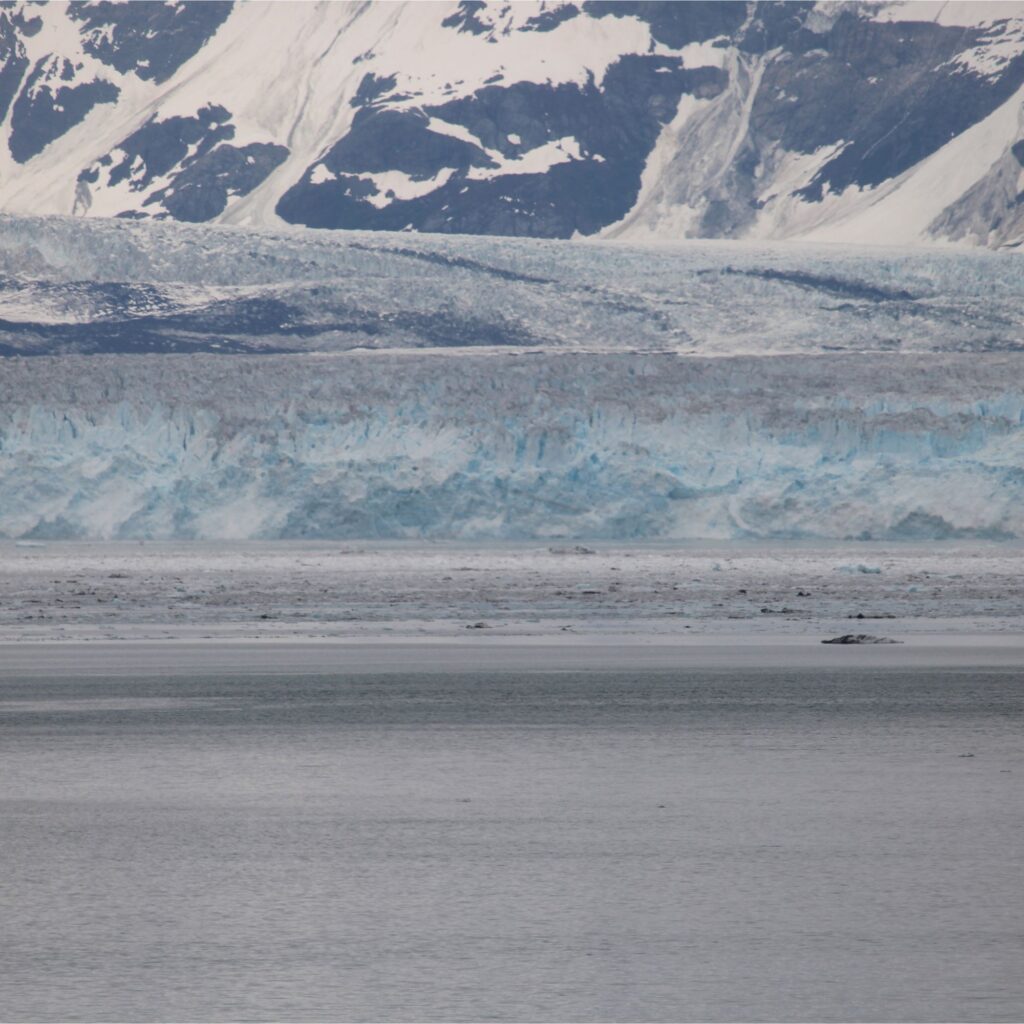 A vast icy landscape featuring a glacier with blue hues, surrounded by snow-capped mountains and a calm body of water in the foreground.