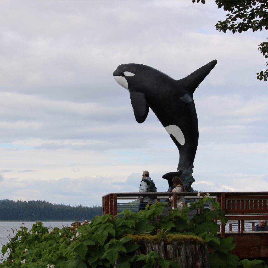 A person standing on a rock sculpture of an orca whale by a waterfront with trees in the background.
