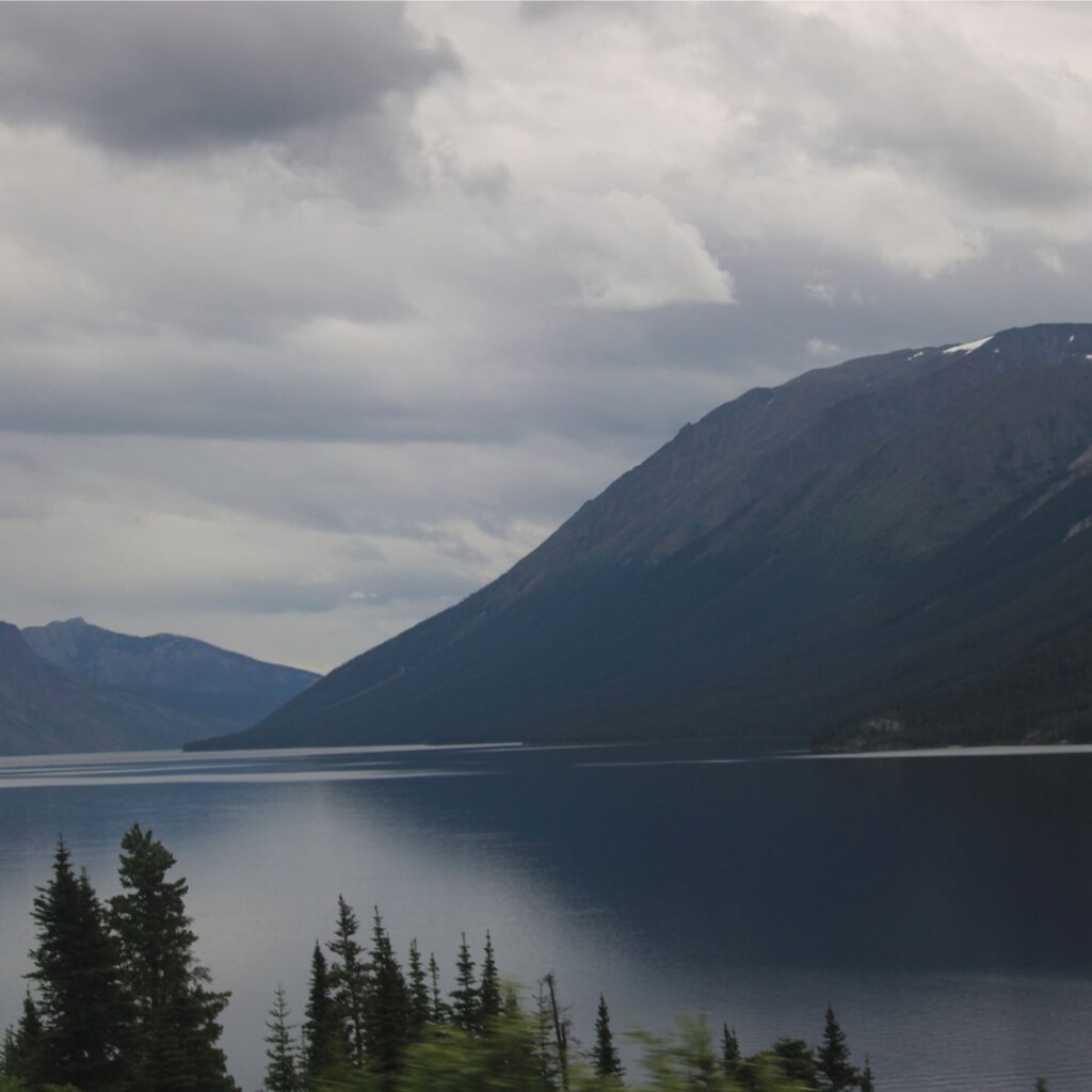 A serene landscape featuring a calm lake surrounded by mountains under cloudy skies.

