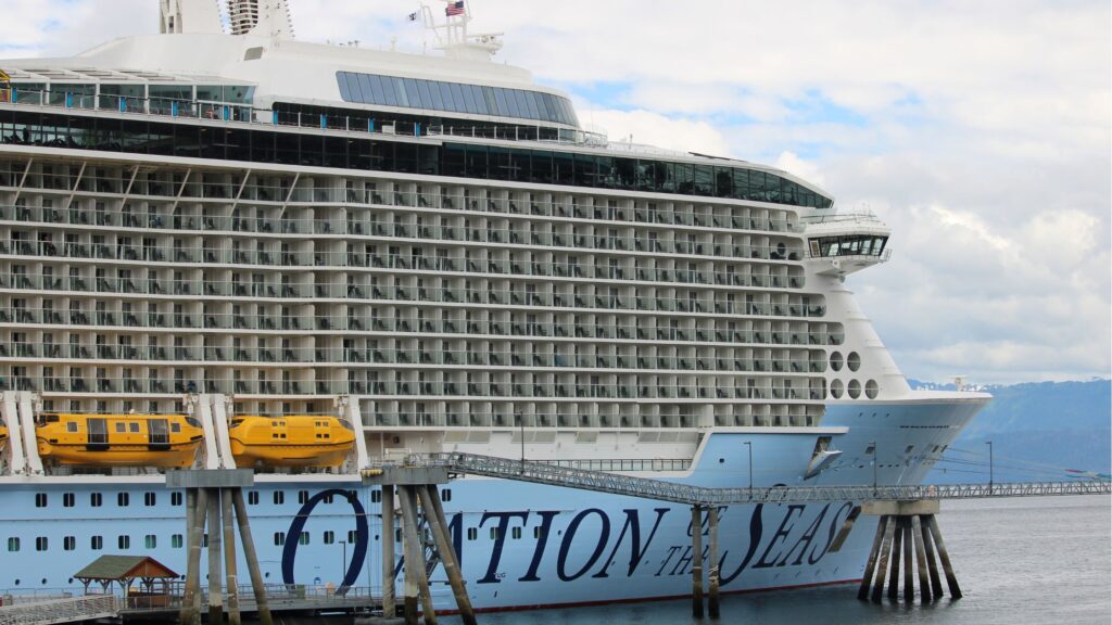A large cruise ship docked in a harbor with the name Ovation of the Seas.