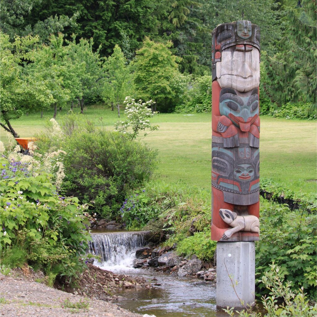 A small, carved wooden totem pole featuring stacked figures painted in red, green, and black, standing next to a small stream and surrounded by trees in the background.