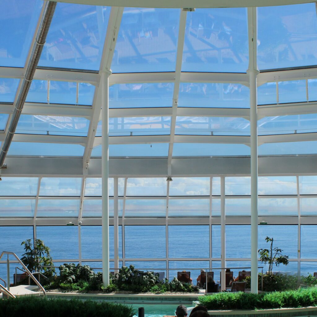 A spacious indoor area with large windows showing a view of the ocean and sky, surrounded by plants.