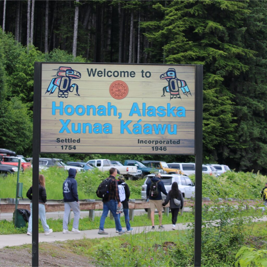 A wood sign among a forested area that reads welcome to Hoonah, Alaska. There are people walking along a sidewalk behind the sign.