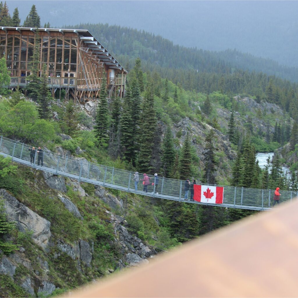 People walking on a suspension bridge, with a Canadian Flag on it, in a forested area, surrounded by rocks and greenery.