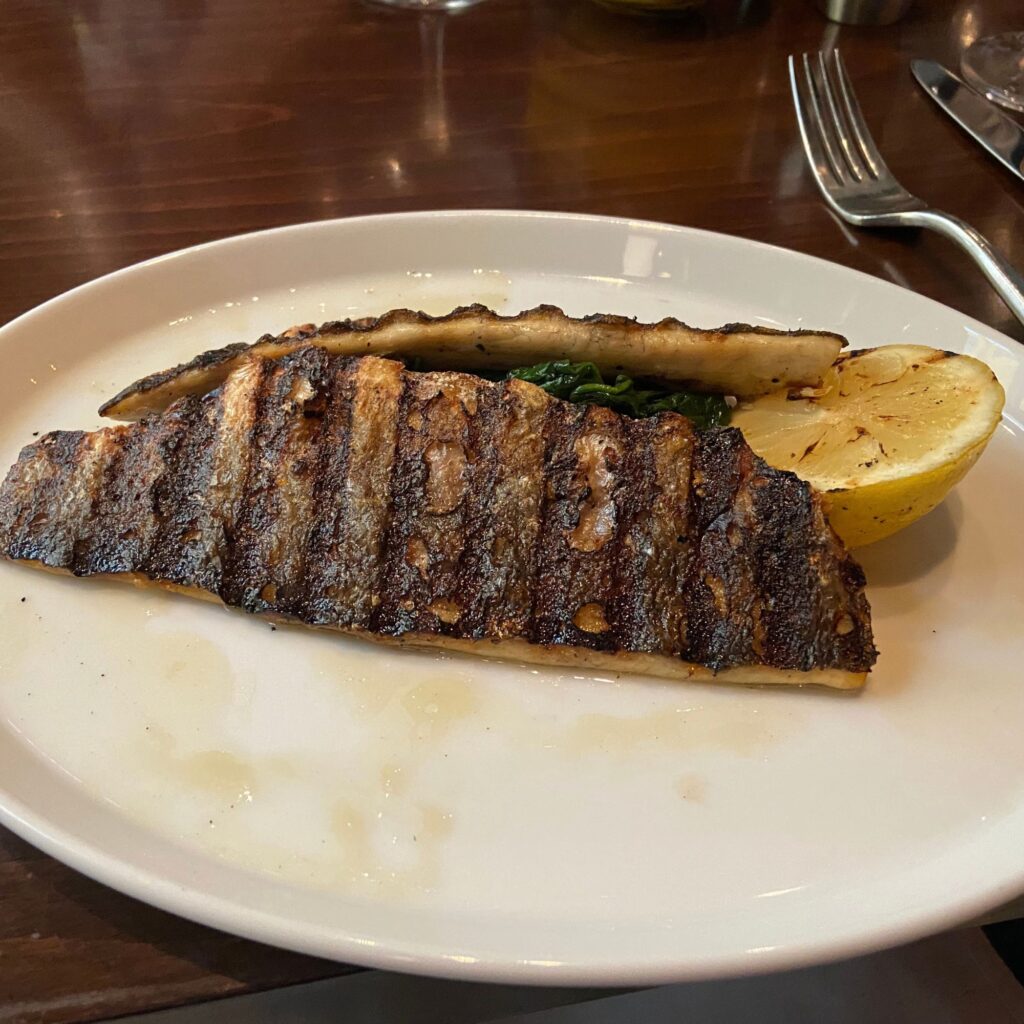 A piece of grilled fish on a white plate with a a half a lemon placed on a wooden table.