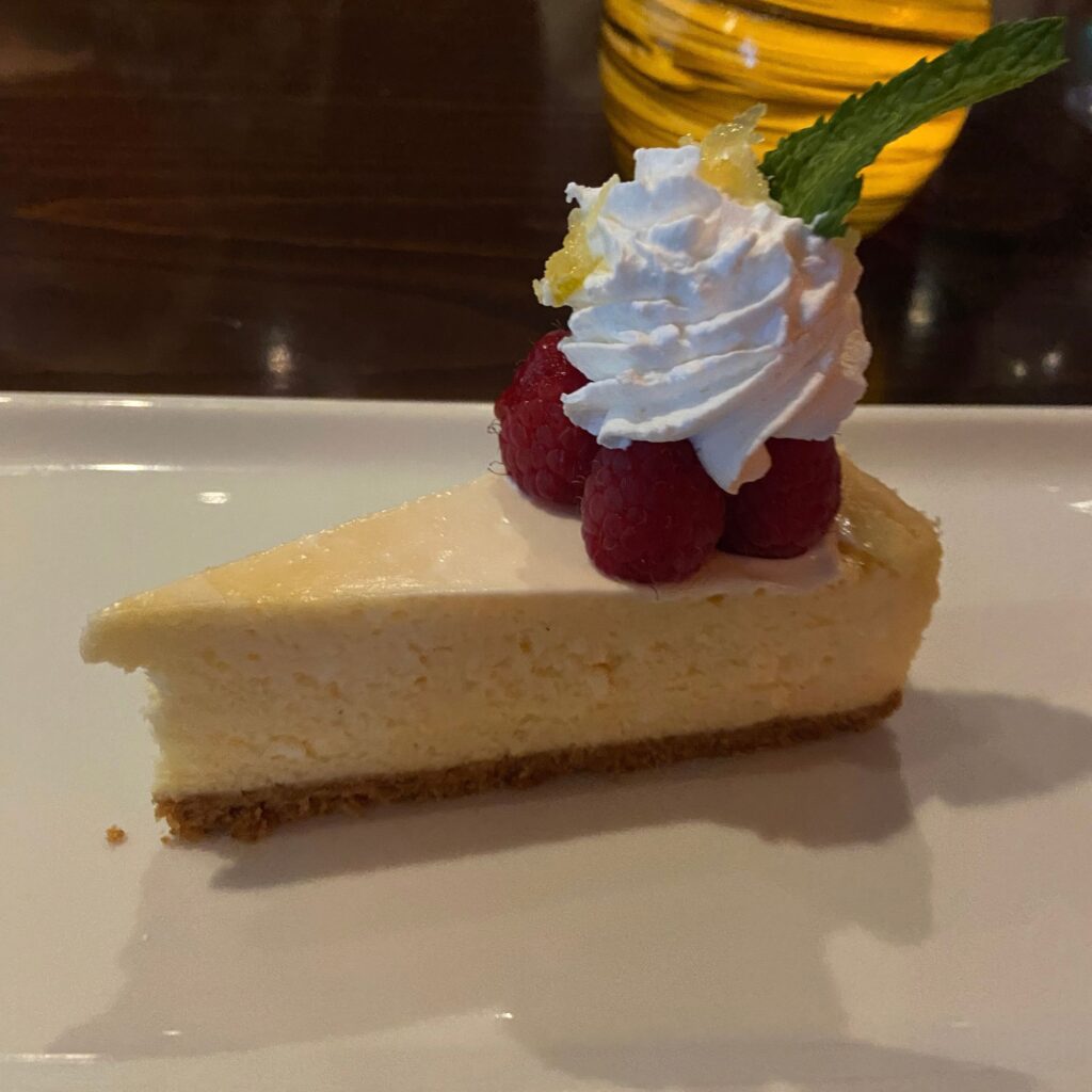 A slice of cheese cake with raspberries, whipped cream and mint leaves on top, served on a white plate.