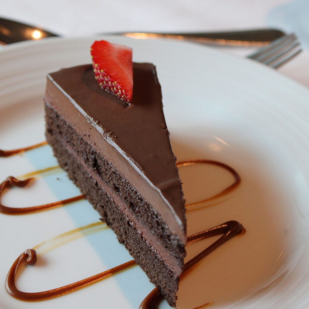 A slice of chocolate cake with a shiny ganache topping and a strawberry slice on top, served on a white plate with decorative sauce drizzles.