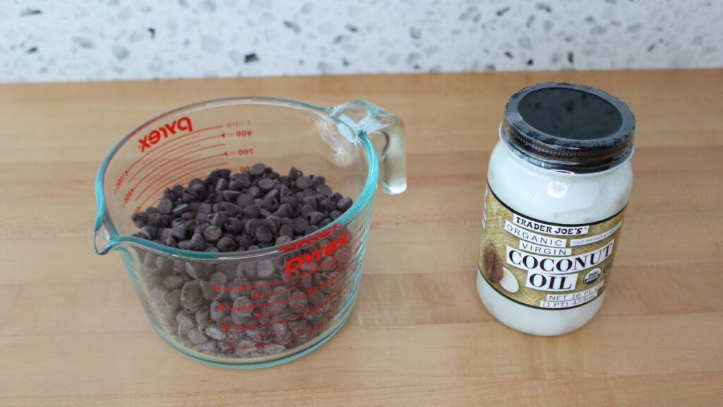 A collection of ingredients chocolate chips in a measuring cup, and a jar of coconut oil, displayed on a wooden countertop.