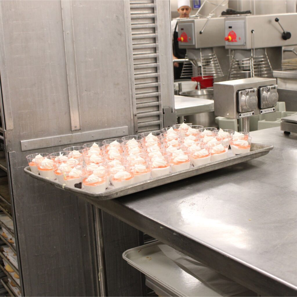 A commercial kitchen with metal countertop containing dessert cups topped with whipped cream.