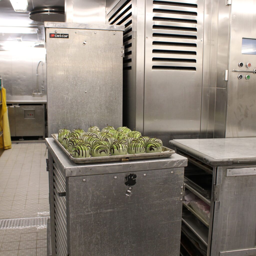 A commercial kitchen with metal shelving and a cart containing dipped whole apples drizzled with chocolate.