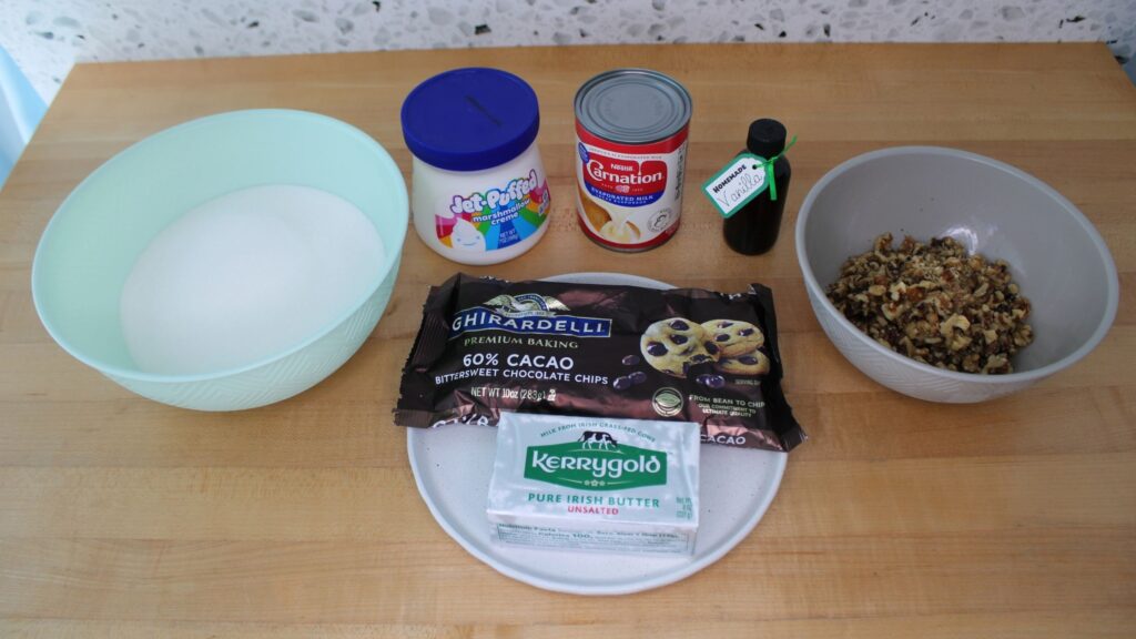 A collection of ingredients including chocolate chips, butter, a bowl of sugar, marshmallow cream, a can of milk, vanilla, a bowl of nuts, displayed on a wooden countertop.