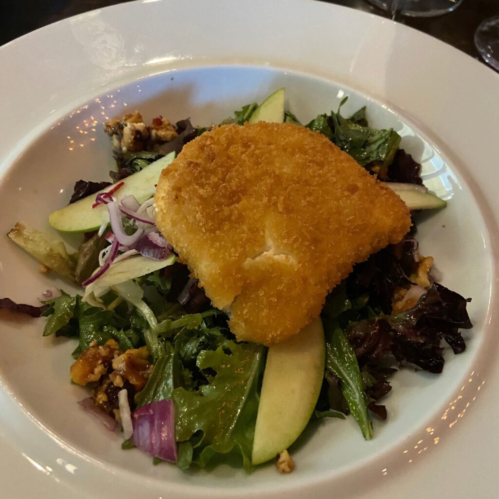 A plate of salad topped with a crispy fried piece of cheese, garnished with apple slices and nuts.