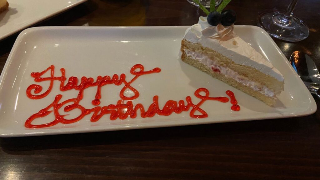 A slice of vanilla cake with strawberry cream filling, topped with whipped cream frosting and blueberries, served on a white plate with red gel writing that says "Happy Birthday!"