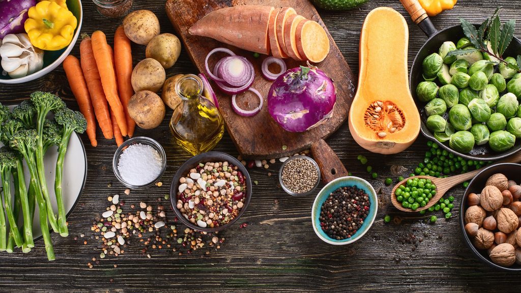 A variety of fresh vegetables and herbs arranged on a wooden surface, including broccoli, sweet potatoes, carrots, potatoes, onions, Brussels sprouts, butternut squash, peas, and several bowls of spices, grains, and a bottle of oil.