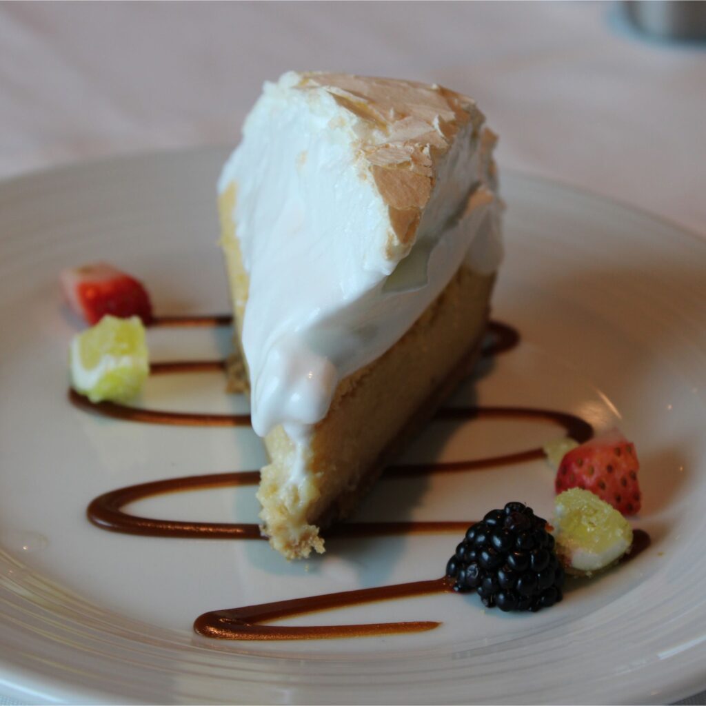 A slice of pie with meringue on top, served on a white plate with decorative sauce drizzles and berries and limes.