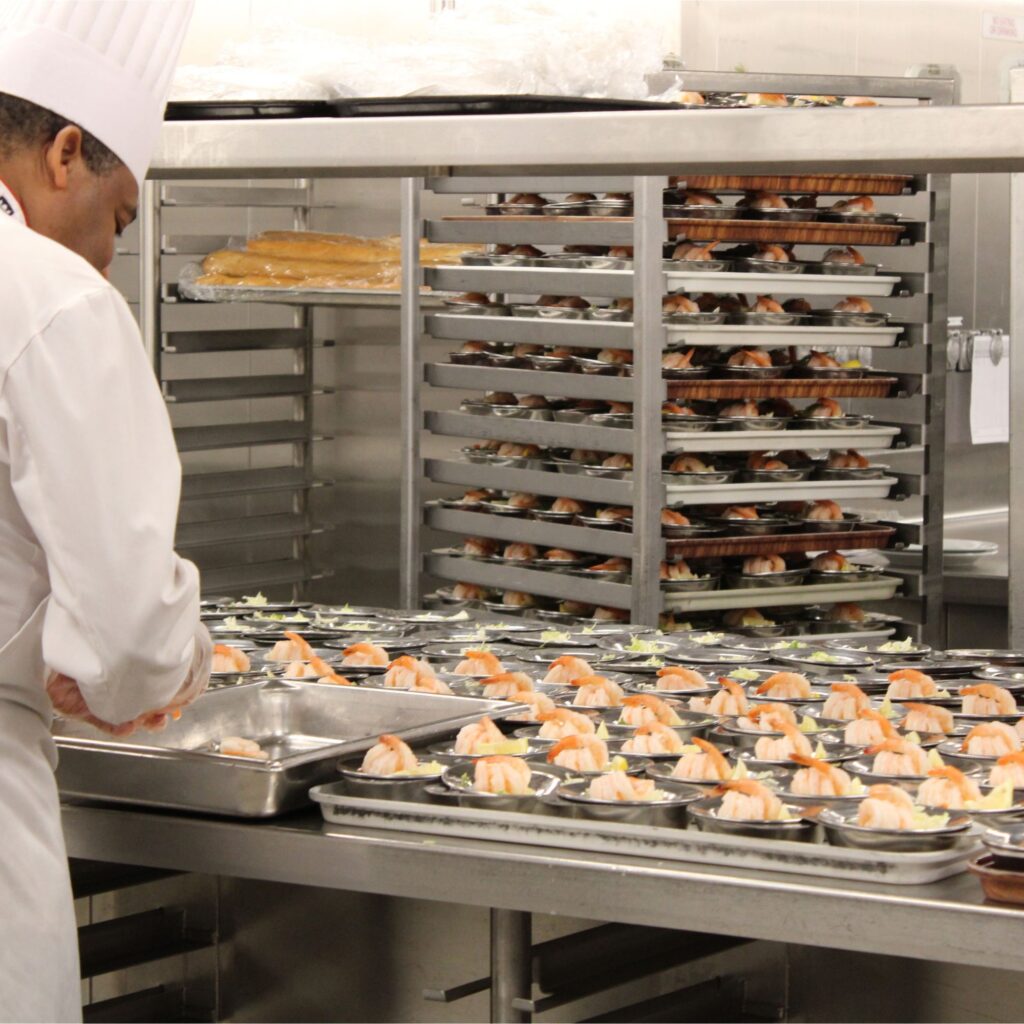 A chef in a kitchen preparing dishes, with trays of food arranged on a table and a rack of more trays in the background.