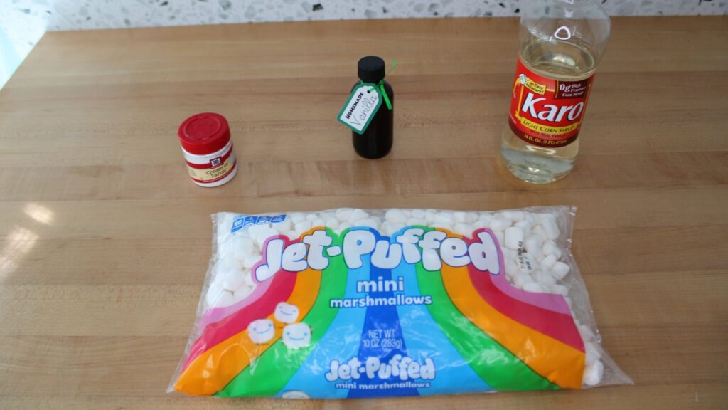 A collection of baking ingredients including marshmallows, cream of tartar, vanilla extract, and Karo light corn syrup, displayed on a wooden countertop.