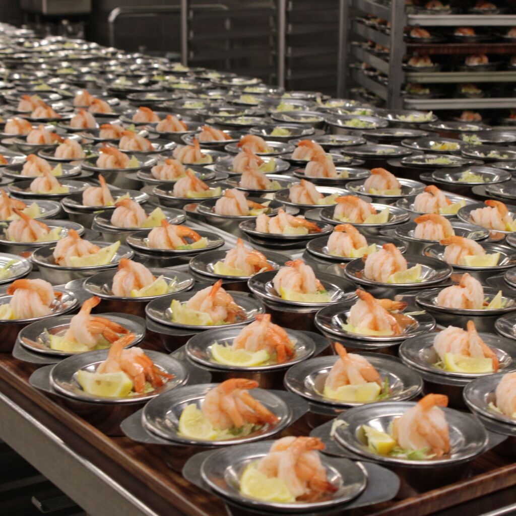 A commercial kitchen with metal countertop containing dozens of bowls containing shrimp and lemon wedges.
