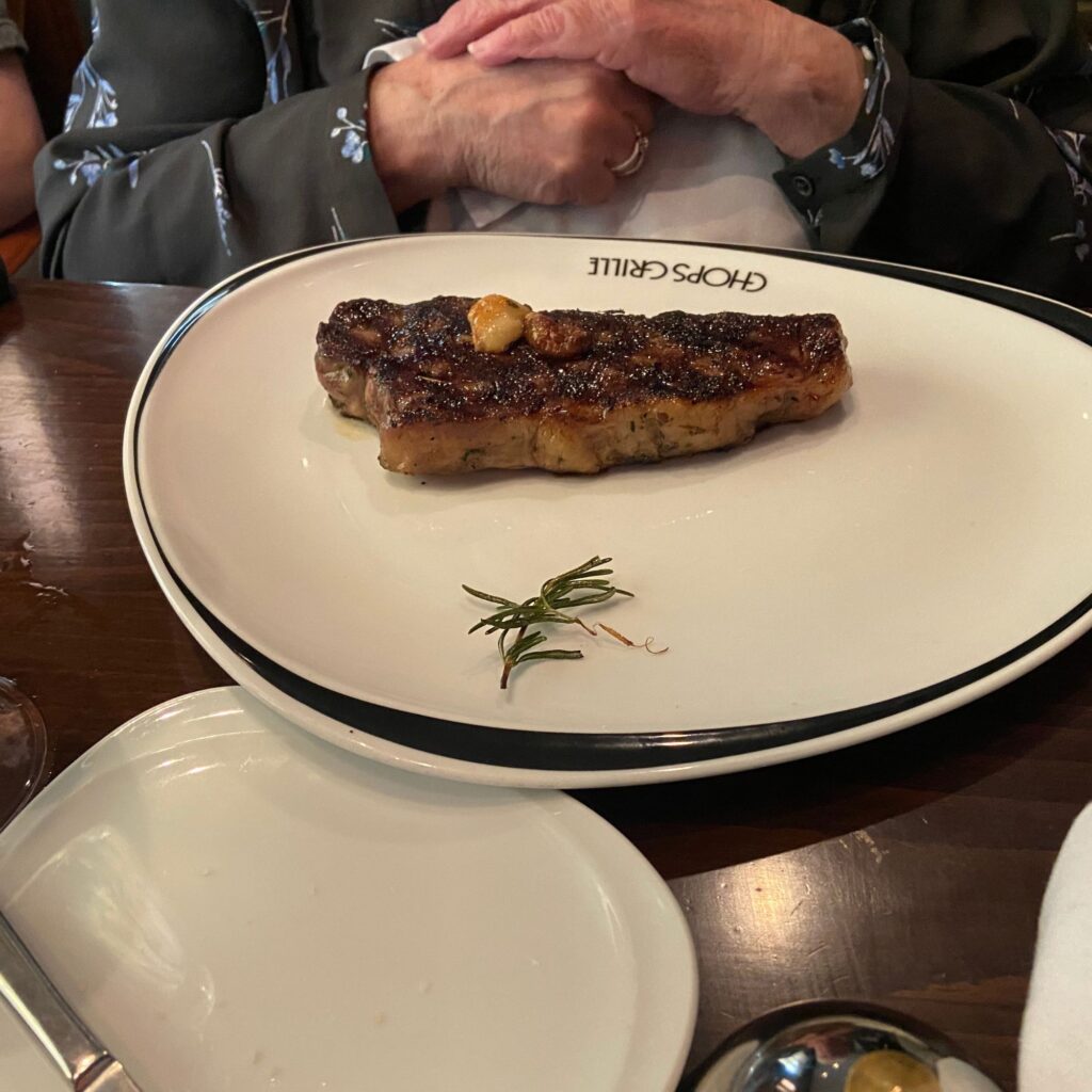 A piece of grilled meat on a white plate with a sprig of herbs, placed on a wooden table.